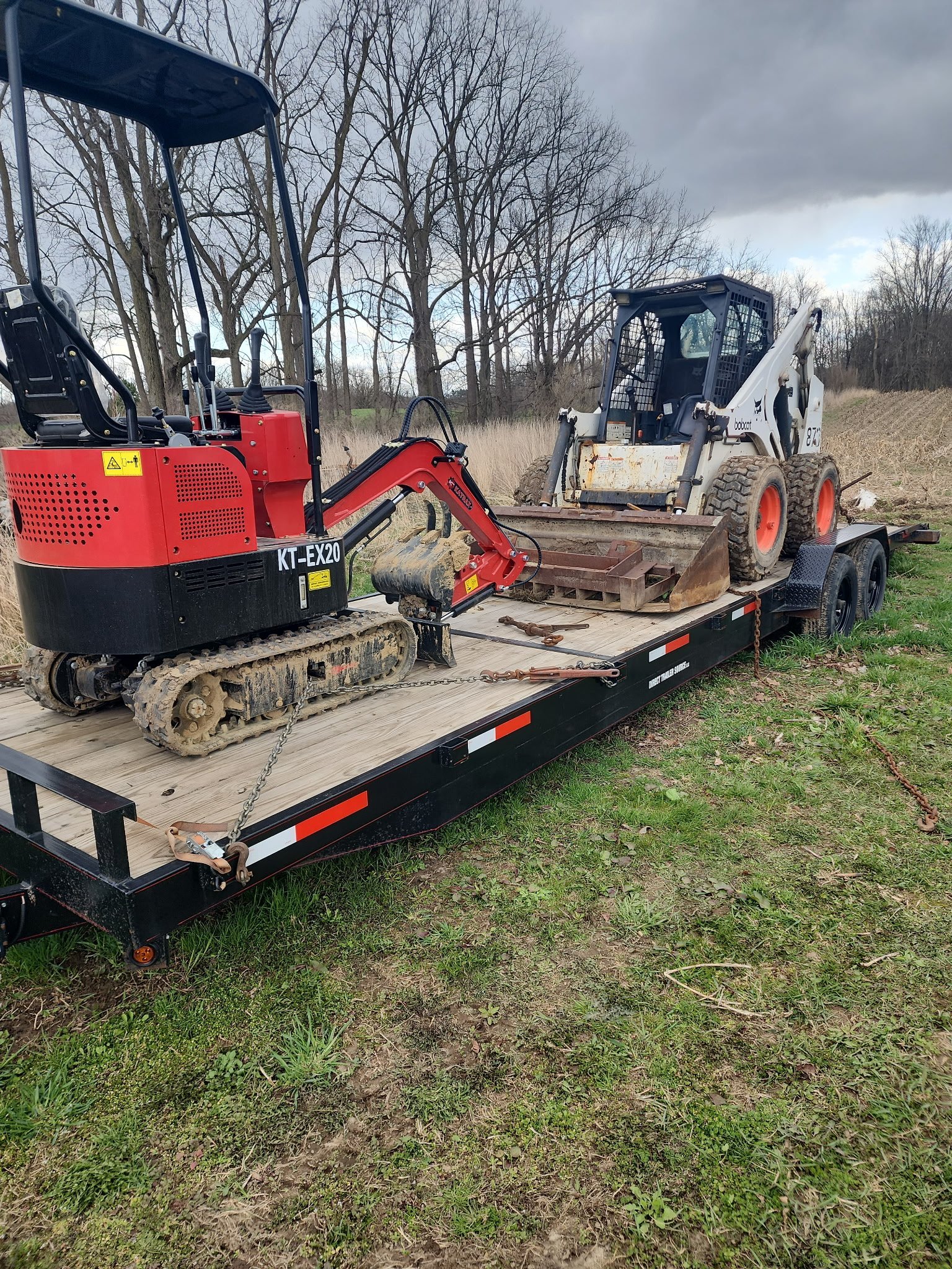 Red compact excavator loaded on a flatbed trailer with a skid steer loader nearby in a rural outdoor setting