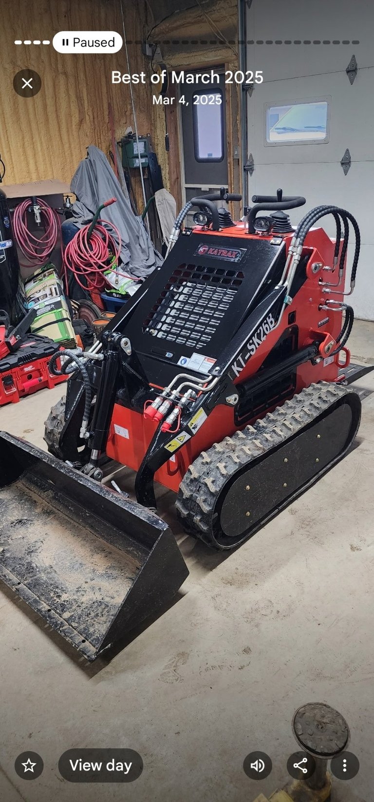 Red and black Komatsu compact track loader in a garage with coiled hoses and equipment on the wooden wall behind it