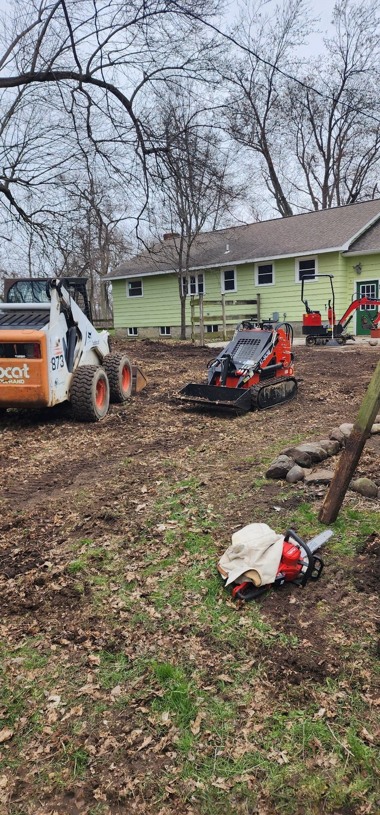 Construction equipment including bobcat and skid steer loader on dirt yard next to green house with bare trees