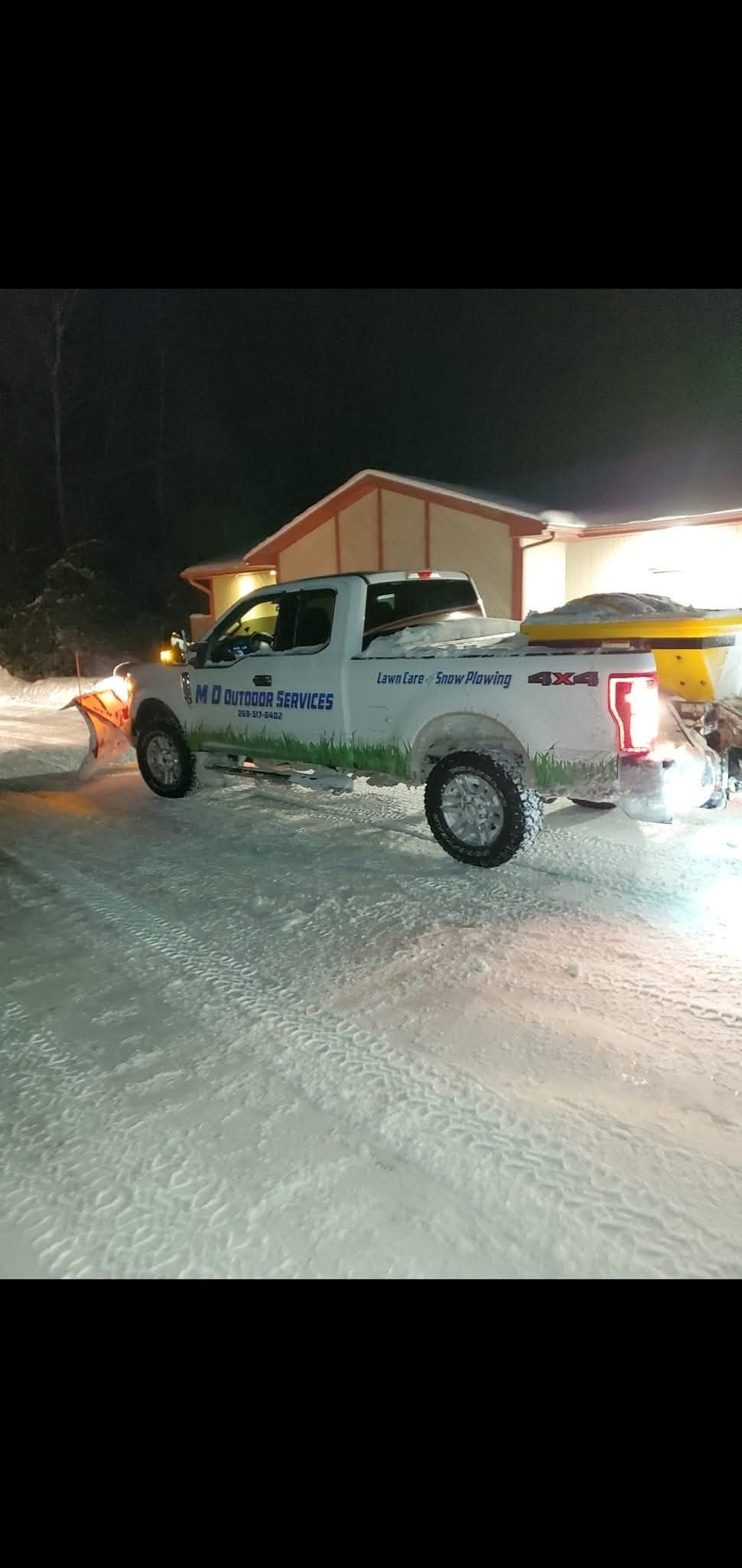 White pickup truck with snow plow attachment parked in snowy driveway at night near illuminated garage